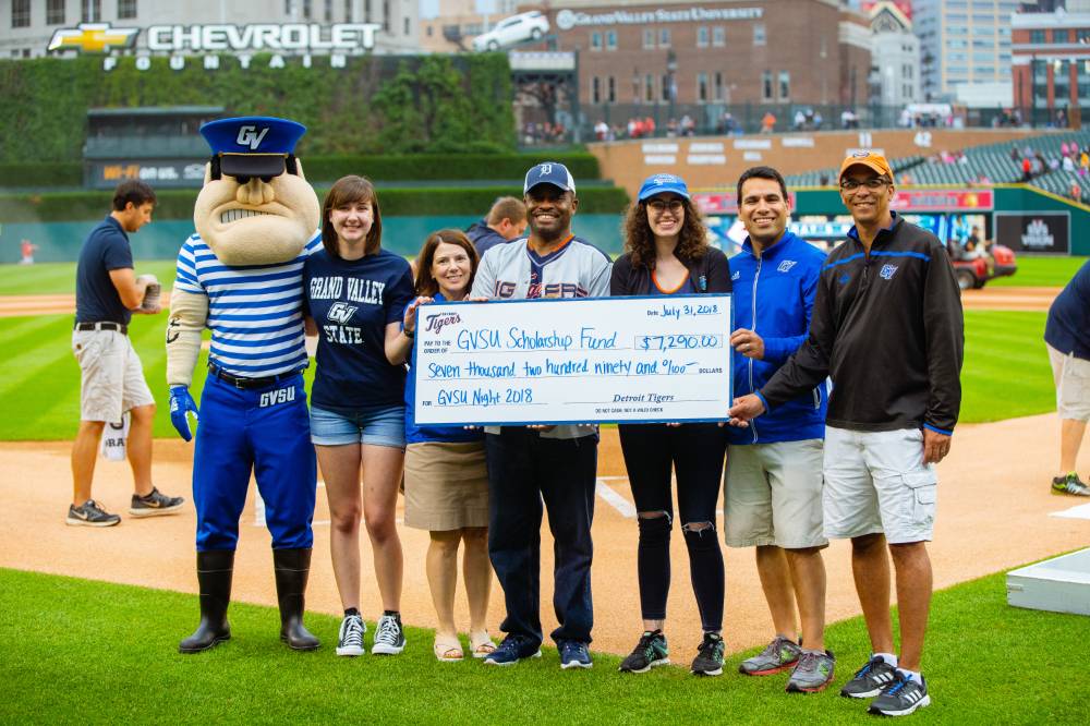 A group of gvsu alumni staff is standing on the infield with louie the laker and a large check that is going into a gvsu scholarship fund.